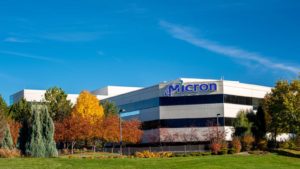 An image of the front of a three story building with the blue logo for "Micron" with grass and trees in front on a clear sunny day.