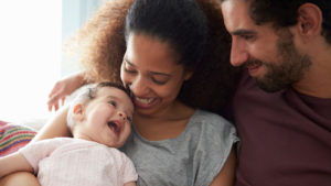 Two smiling parents hold a smiling baby while sitting down.