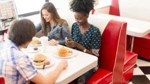 Three young adult friends sit around a vintage restaurant booth eating hamburgers. image represents restaurant stocks to buy