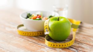 a bowl of vegetables, a green apple, and a measuring tape sit on a wooden table