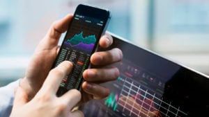 A person holds a phone with a stock chart visible on it with another chart visible on a computer nearby. Best Stocks to Buy