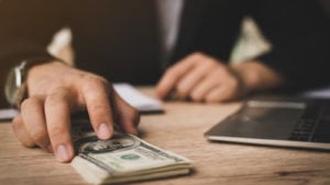 hands at desk near laptop computer, with one hand holding a pile of hundred dollar bills. Bank stocks