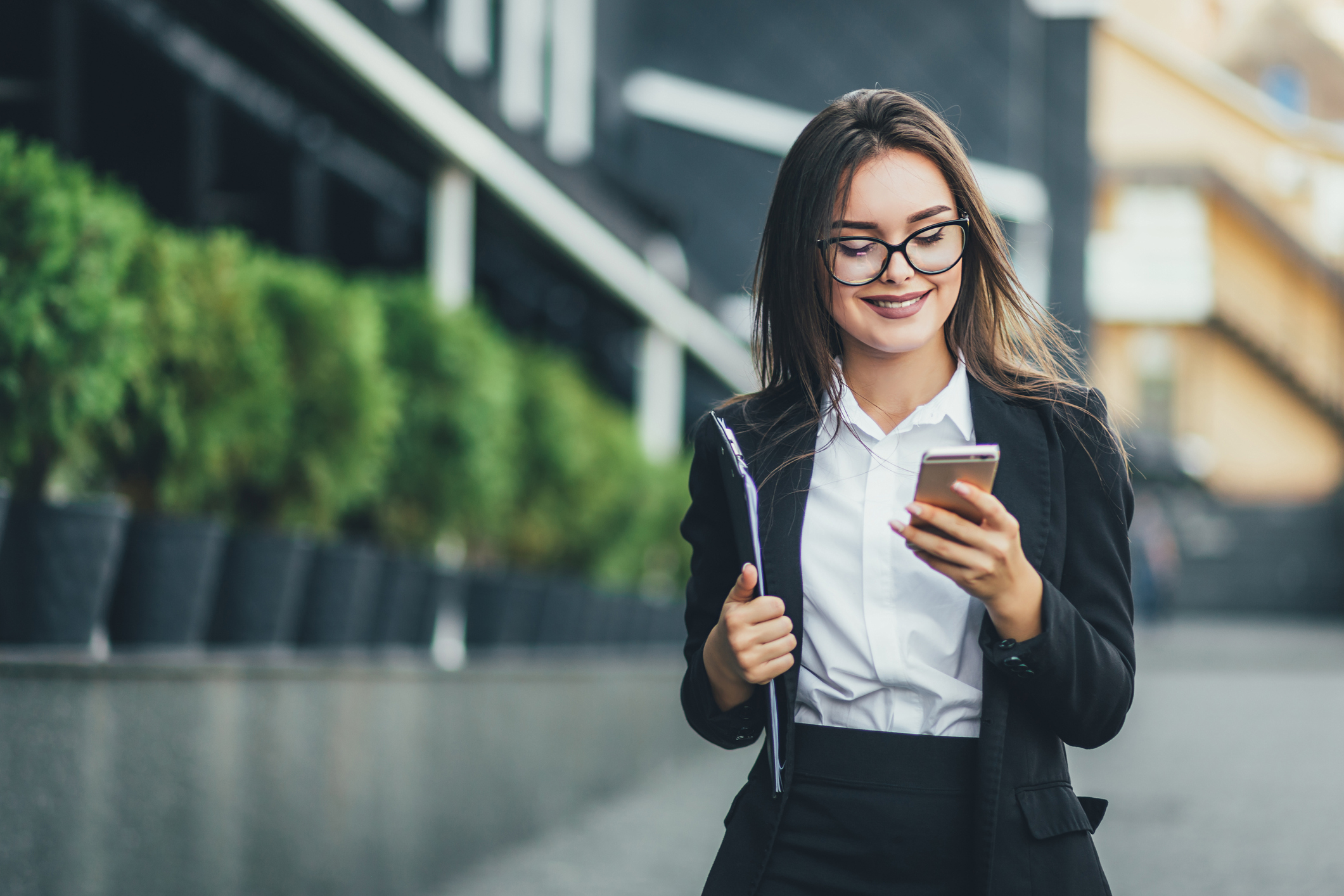 An investor walks outdoors in a city and looks at something on a phone.