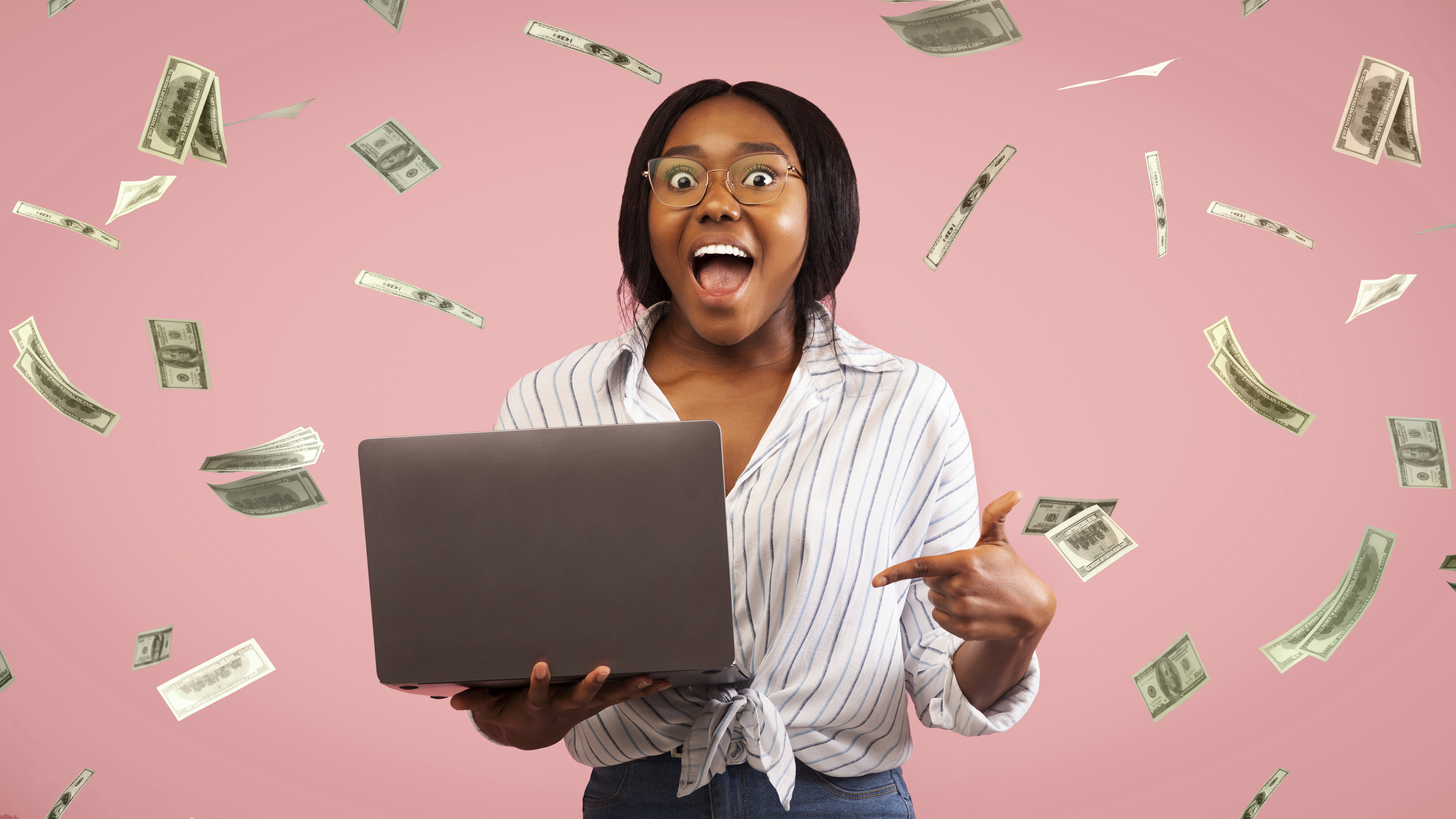 A person with mouth open and eyes wide, holding a laptop as paper money falls around them. 