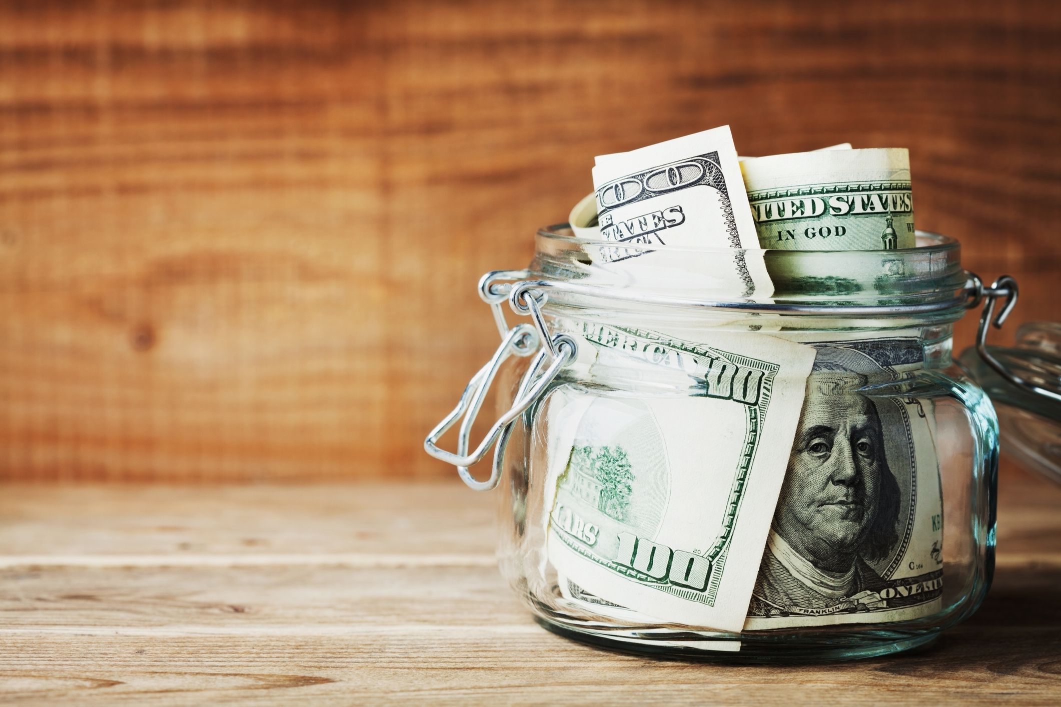 A jar full of $100 bills on a wooden desk.