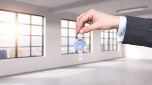 hand of person in a suit dangling keys with a house symbol on the ring. Windows overlooking city skyline in background.