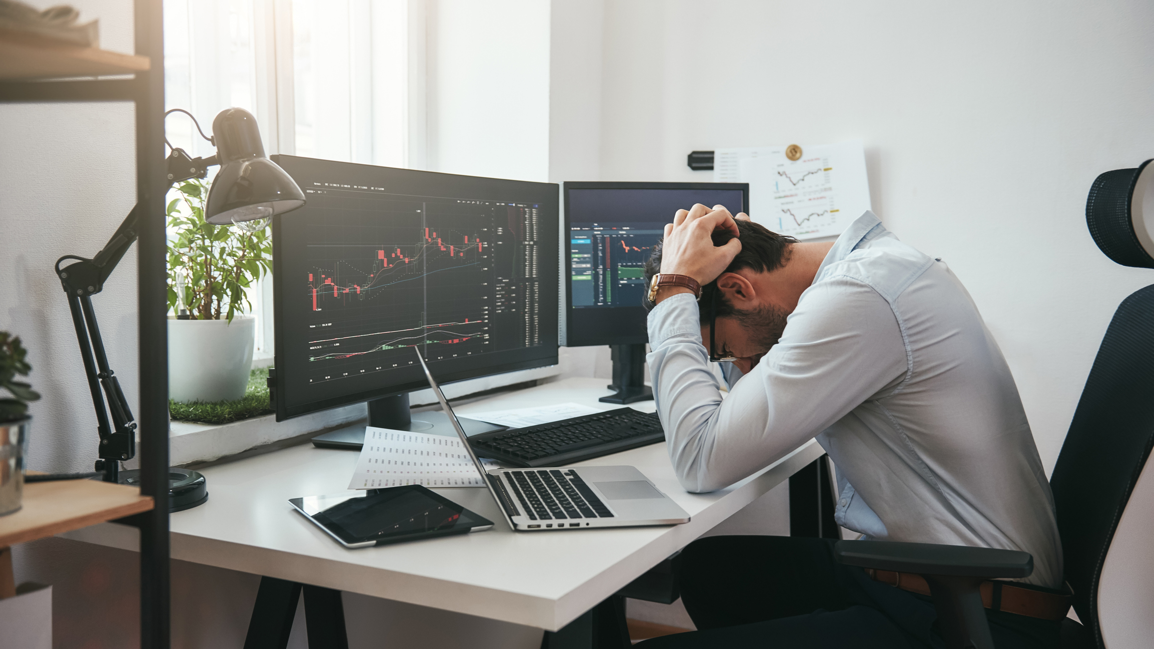 Person in front of computer screens with charts, holding head.