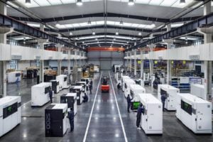 A picture of multiple 3D Printers with people working on them in a factory.