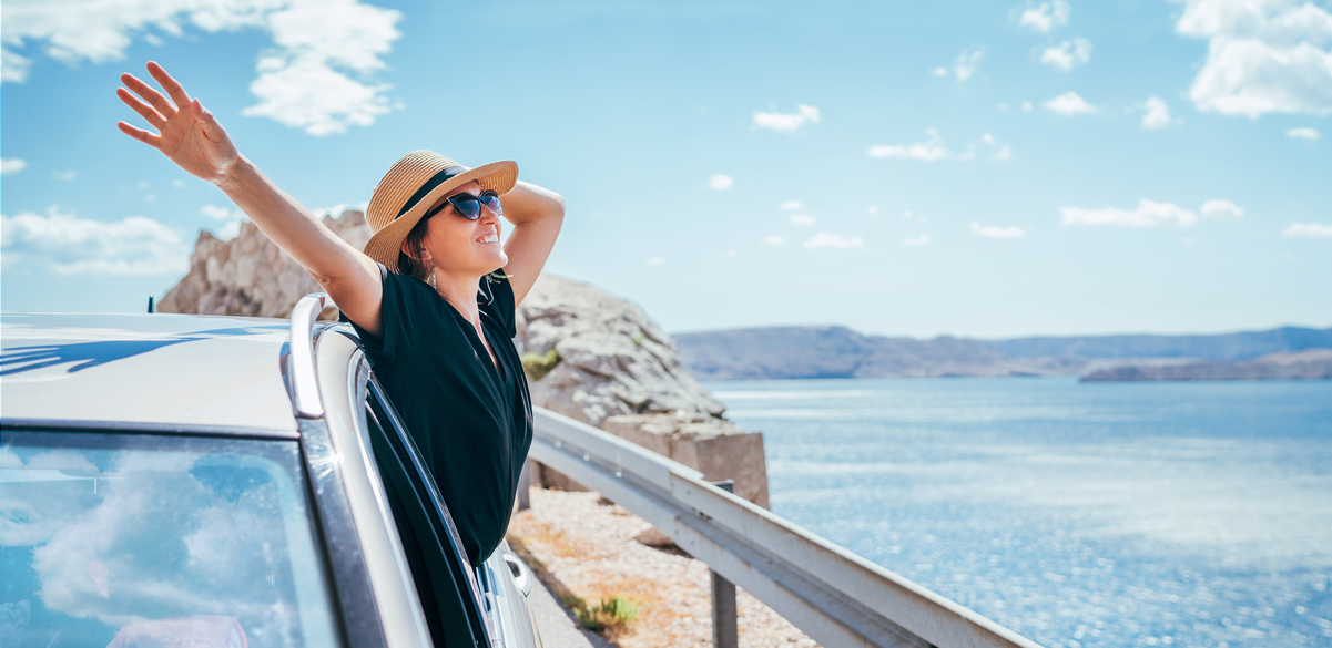 Smiling person with outstretched arms leaning out a car window by a body of water.