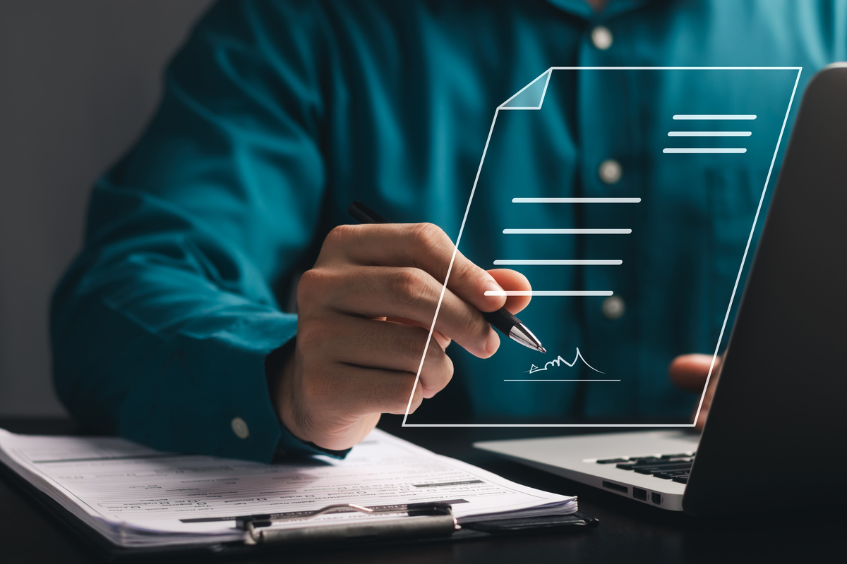 A person in a dress shirt signing a document that's displayed digitally above their desk.