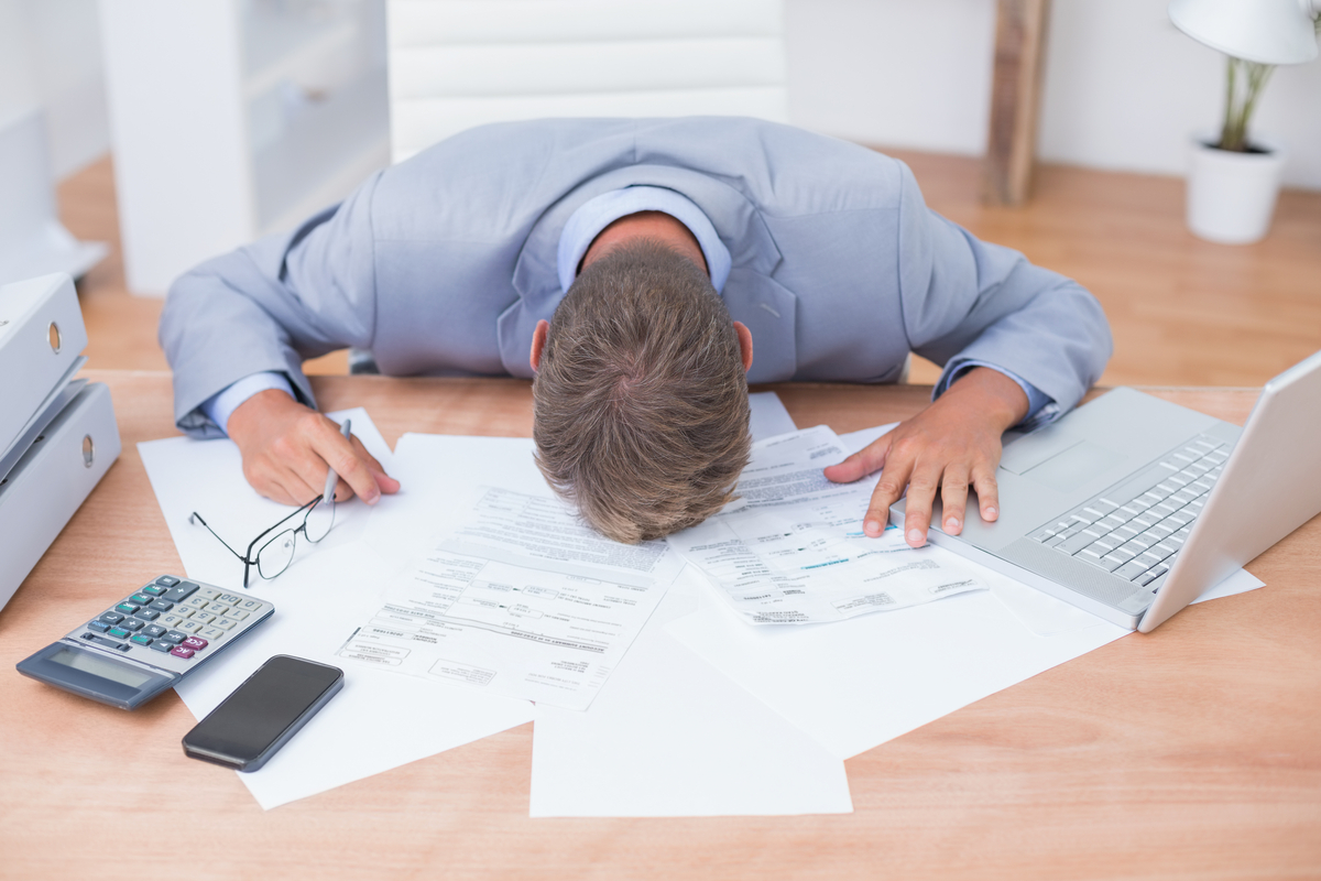 Frustrated business person with their head down on a desk on top of a pile of financial documents, next to a computer and calculator.