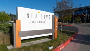 An image of a white, grey, and orange sign with the "Intuitive Surgical" logo and number "1020" on a patch of grass in front of a white, grey, and orange building on a sunny day.