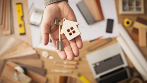 Real estate agent handing over a house key, desktop with tools, wood swatches and computer on background, top view. Real estate stocks.