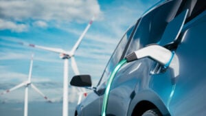 Photo of steel blue electric car being charged with wind silos and blue sky in the background. EV