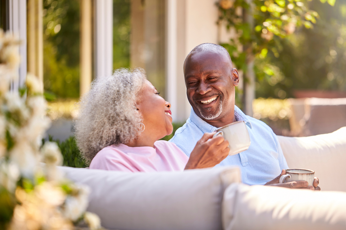 A senior couple smiling at each other while drinking from mugs outdoors.