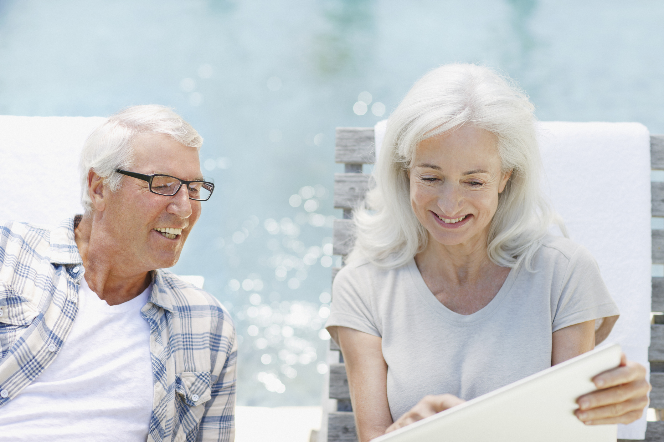 Two people smile and look at something on a tablet by a pool.