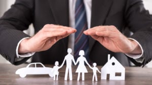 Man in suit with hands over paper cutouts of family, car and home. Represents insurance.