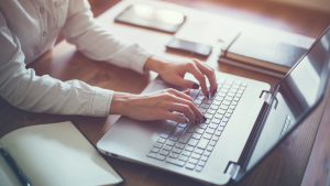 A photo of a person's hands on a laptop keyboard. Several notebooks are on a desk surrounding the laptop.