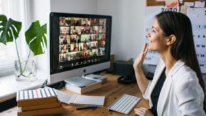 A woman sitting at a desk waves at a large number of people on the videoconferencing software Zoom (ZM).