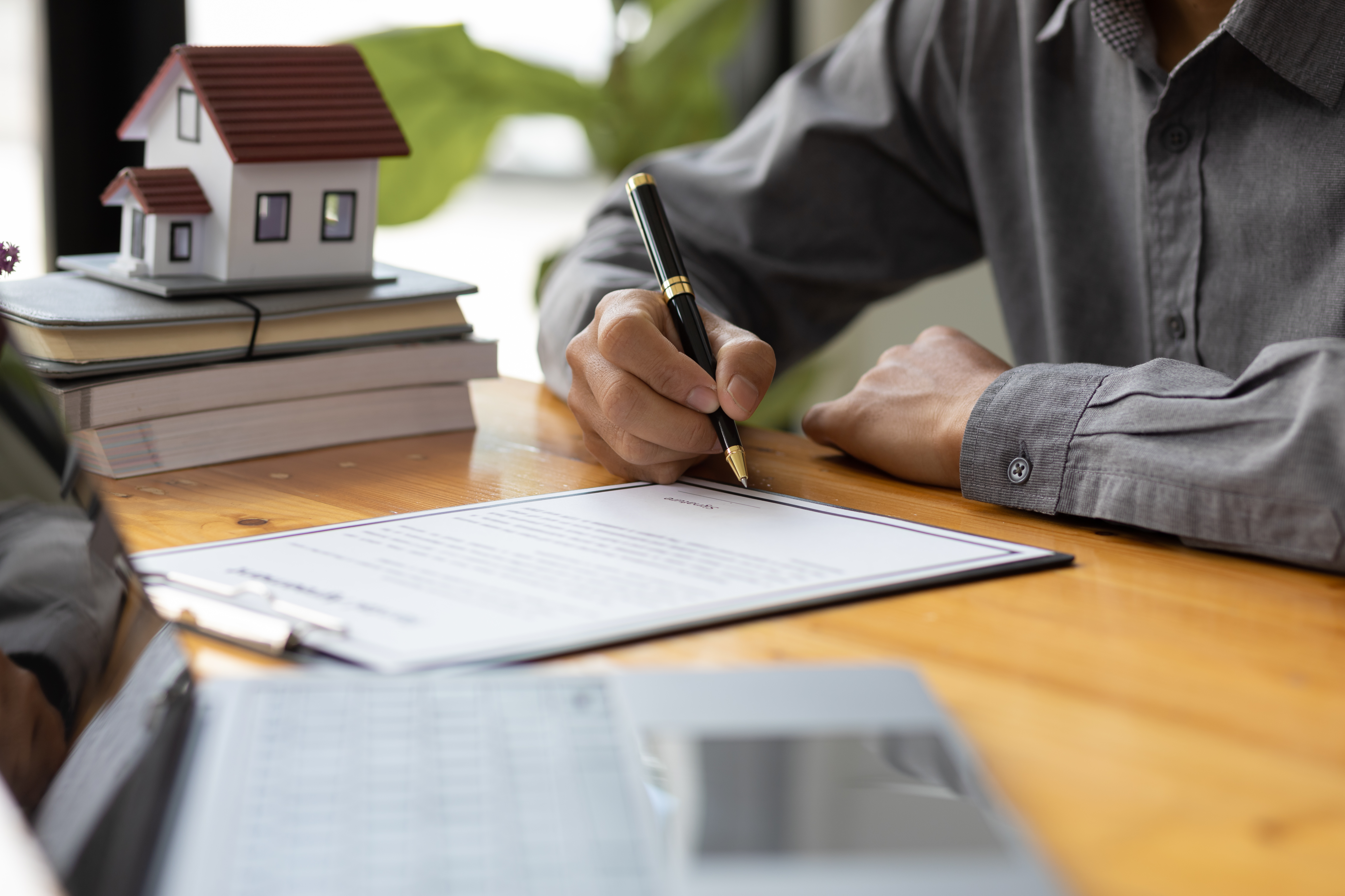 Person signs paperwork with a small home in the background.