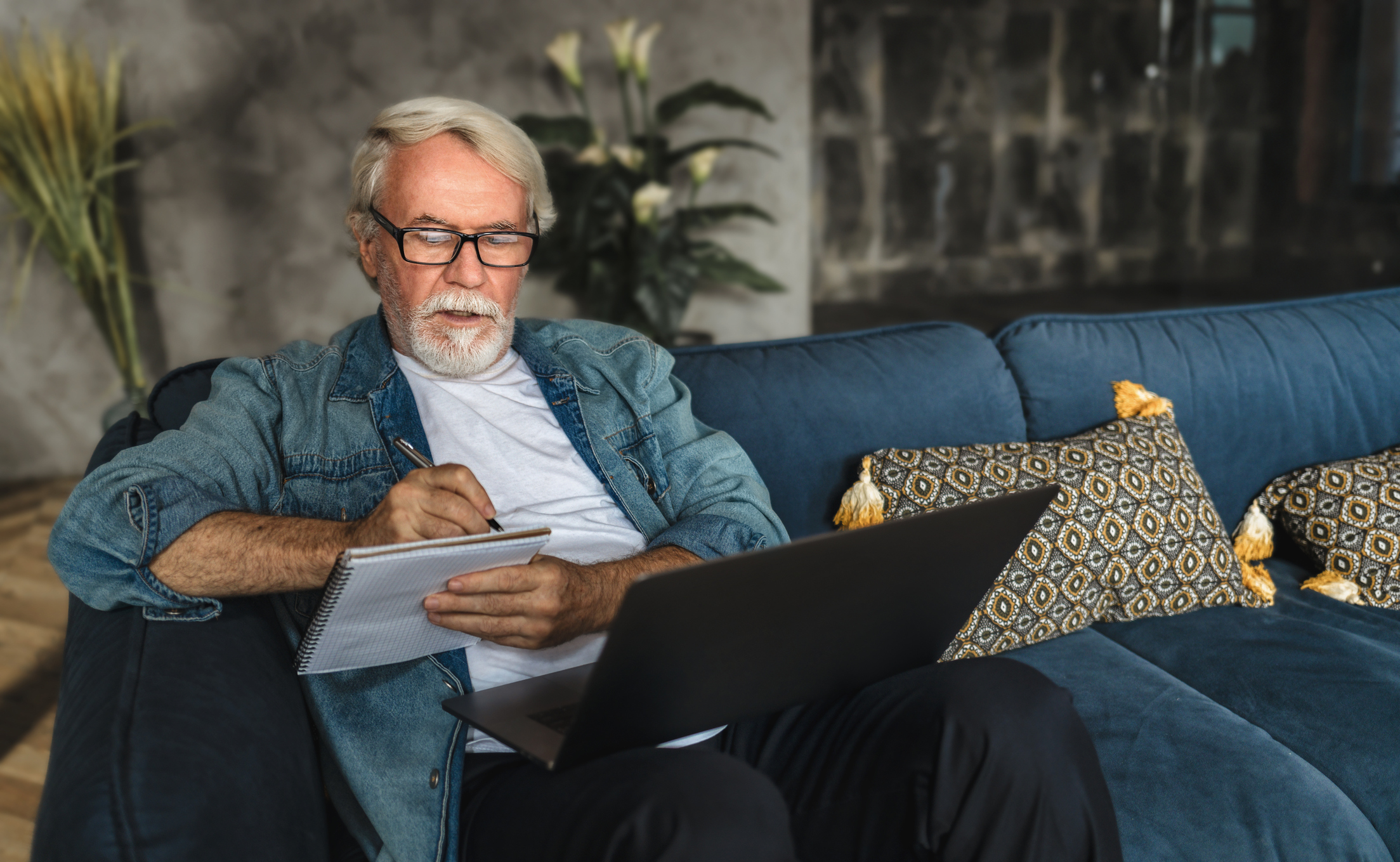 Person writing note in notebook while holding laptop on lap.