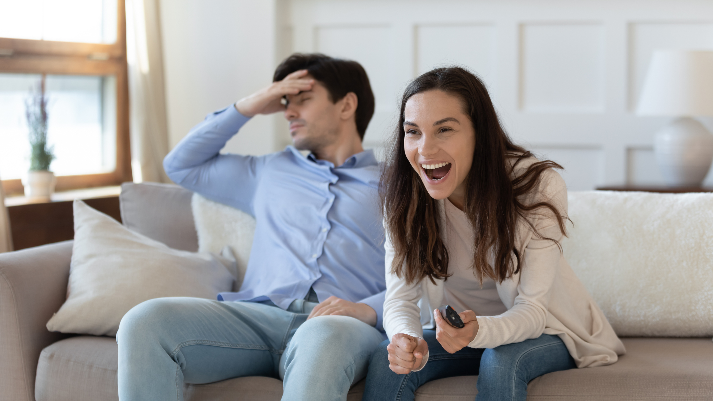 Two people sit on a couch. One is laughing at the TV, and the other is frowning and looking away.