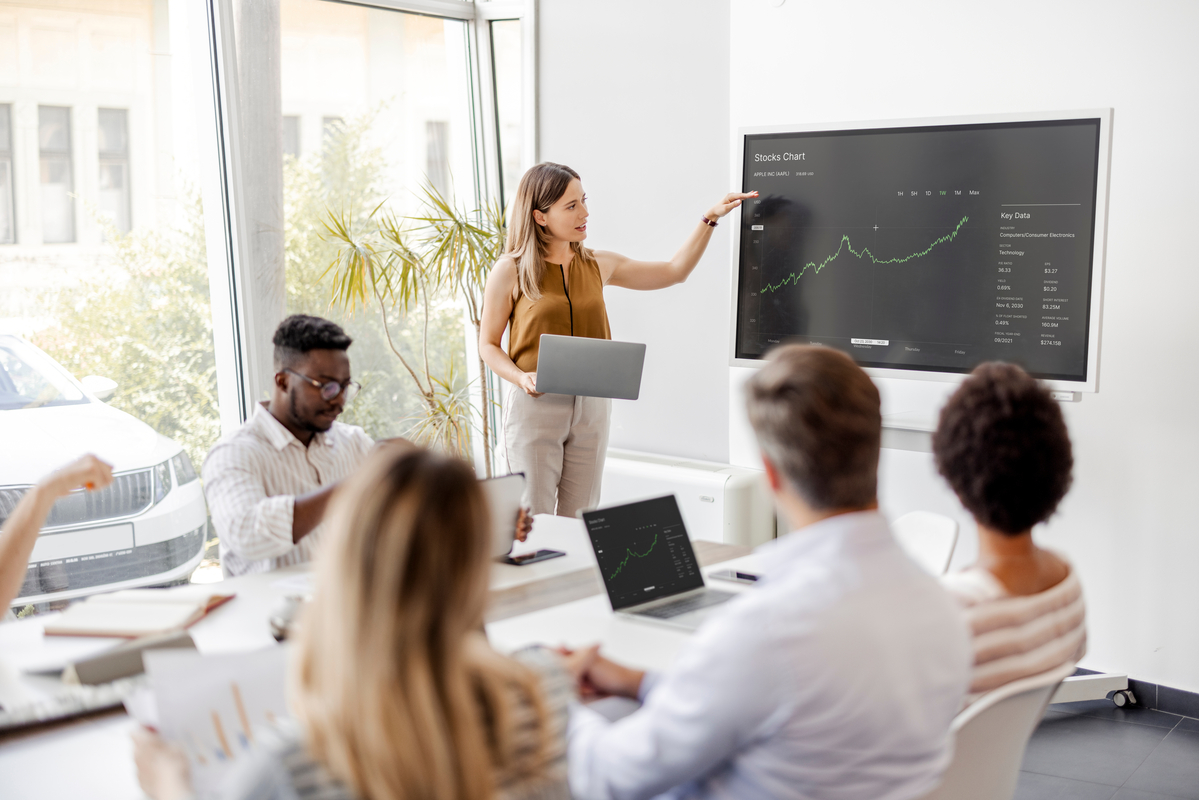 Someone pointing to a stock chart as several people sit at a table.