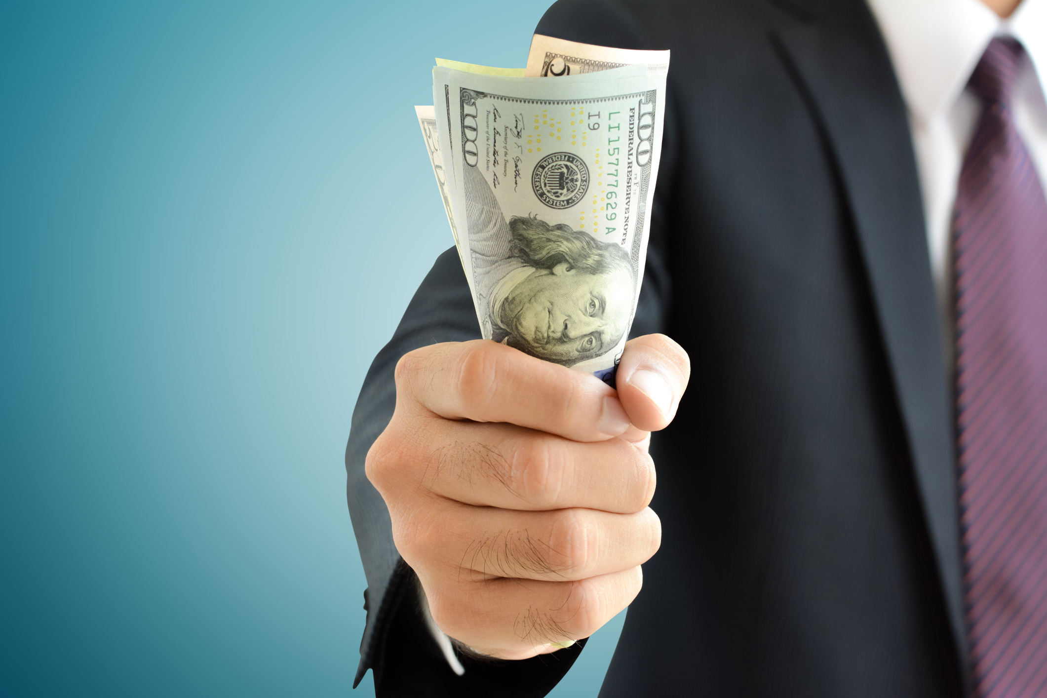 In this close up view of a person in a suit, a hand holds out $100 dollar bills.