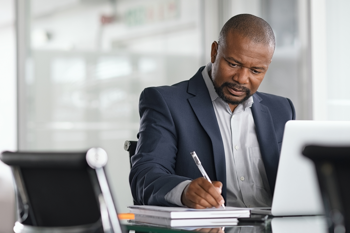 A person dressed in professional attire sitting at a table in front of a laptop and writing with a pen.