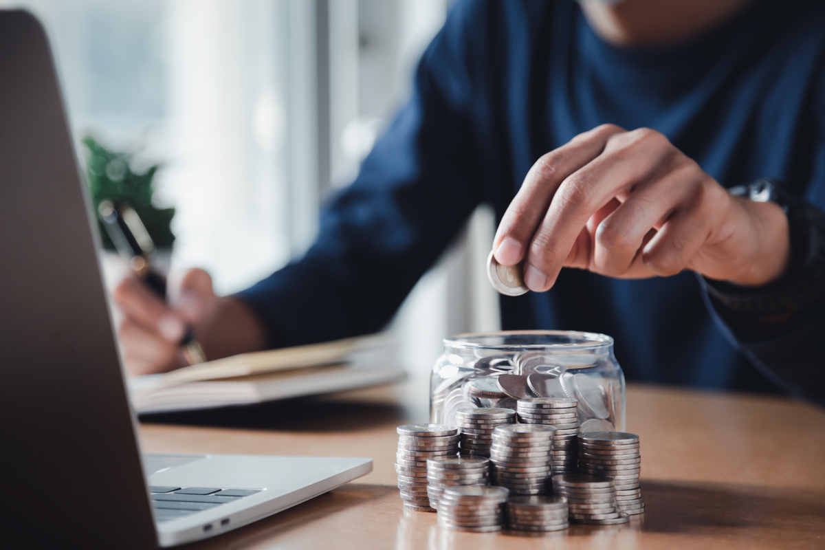 A person stacking coins in a jar while sitting at a table in front of a computer. 