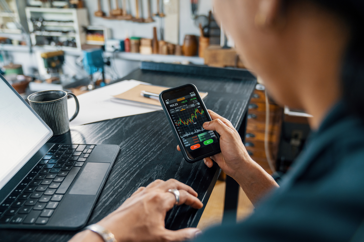 Person at workstation holding smartphone device displaying financial data.