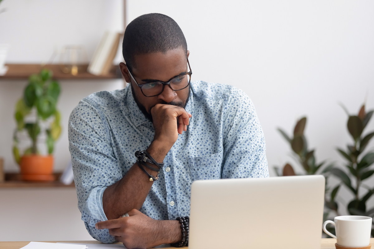 A person sitting down at a table in front of a laptop computer clinches fist and looks seriously at the computer. 