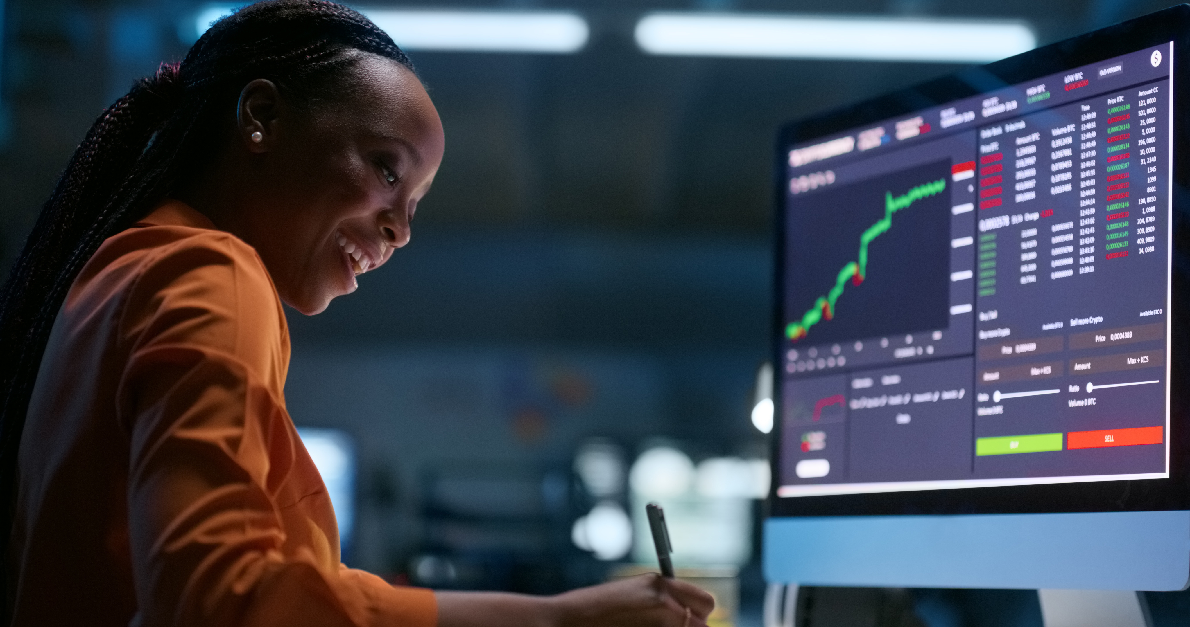 A smiling person writing notes while looking at stock charts on the computer.