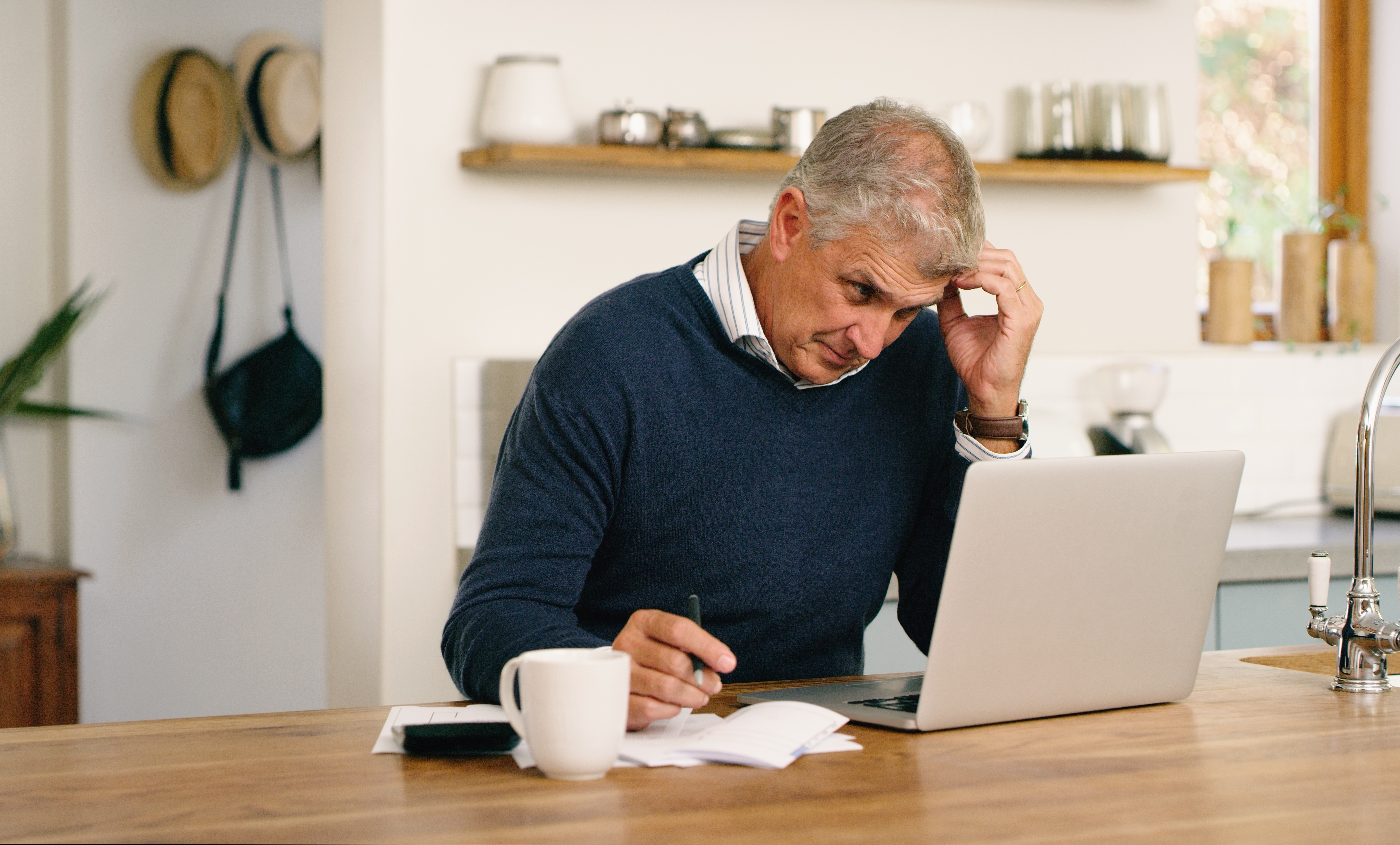 A person looking at a laptop with a pen and paper by his side is taking notes.